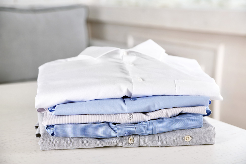 A neatly folded stack of four button-down shirts in white, light blue, blue, and gray on a white table.