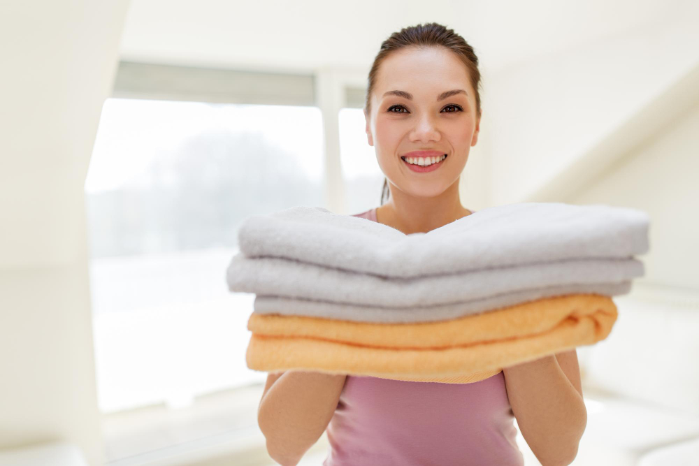 A woman smiles and holds a stack of folded towels, including gray and orange towels, in a brightly lit room.