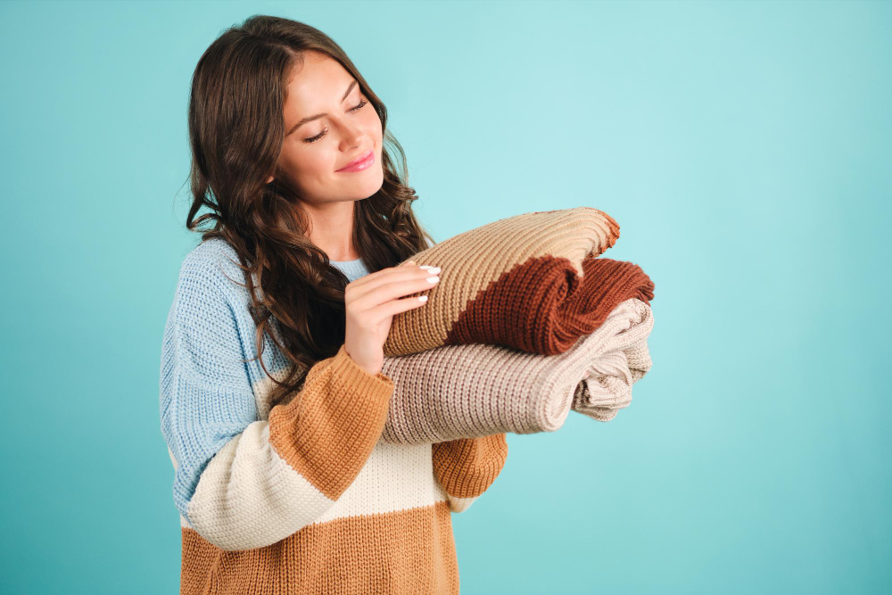 A woman in a striped sweater holds a stack of folded knit blankets against a light blue background.