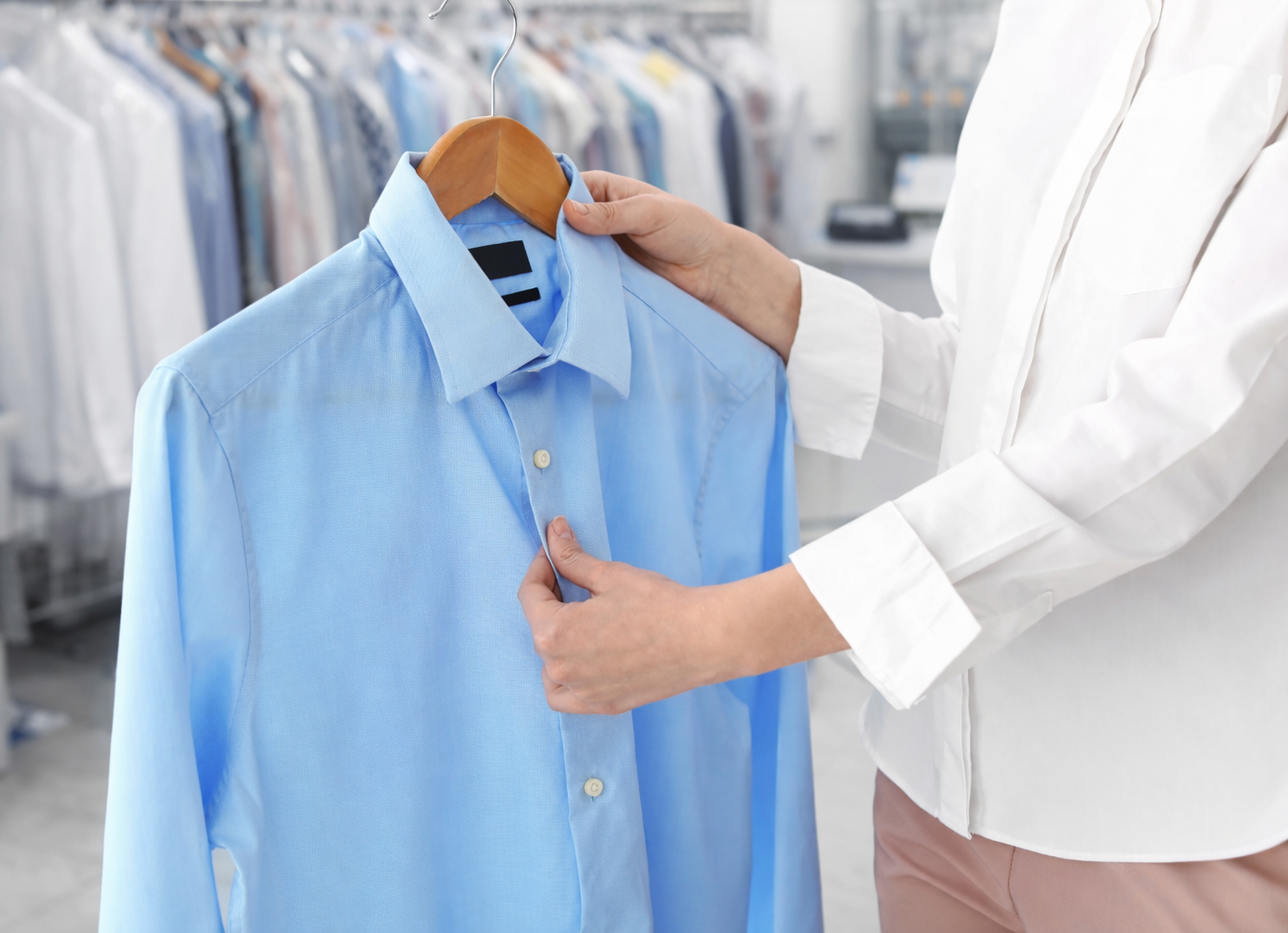 A person holds a light blue dress shirt on a hanger, checking its buttons, with racks of clothes in the background.