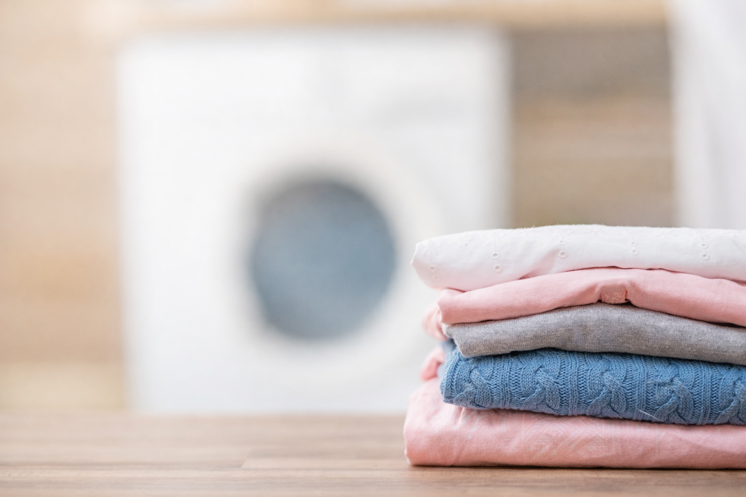 A neatly folded stack of clothes, including white, pink, gray, and blue items, sits on a wooden surface with a washing machine in the blurry background.