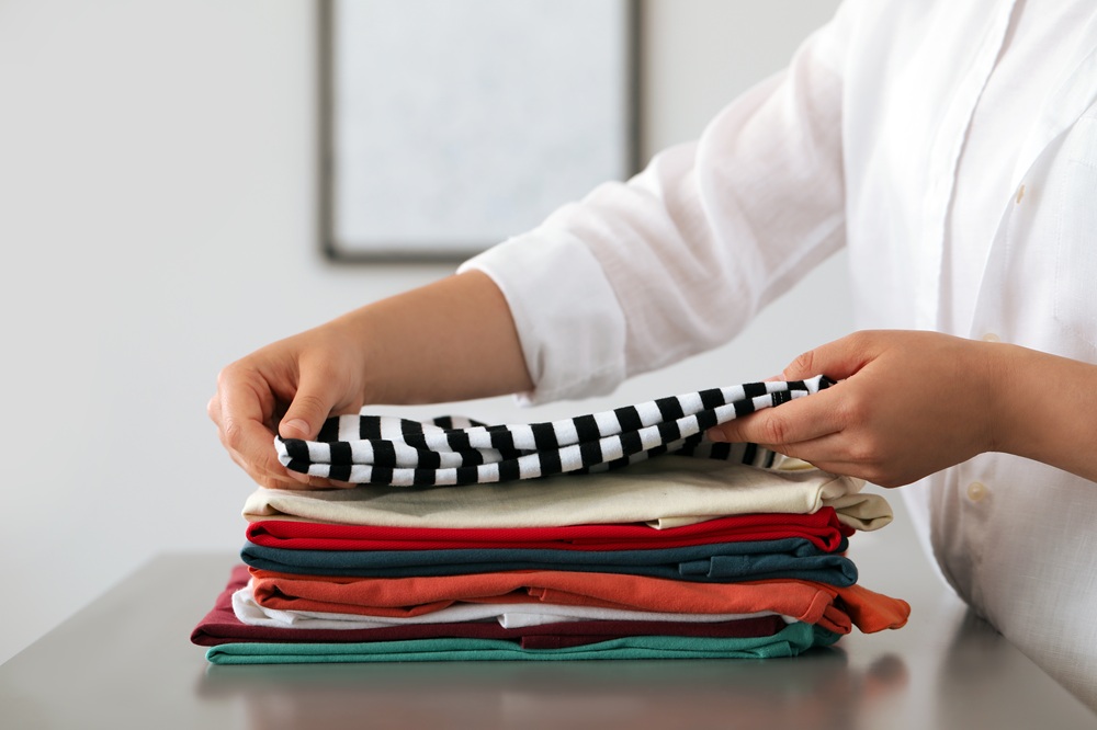 A person folds a stack of neatly arranged clothes on a table, placing a black-and-white striped shirt on top.