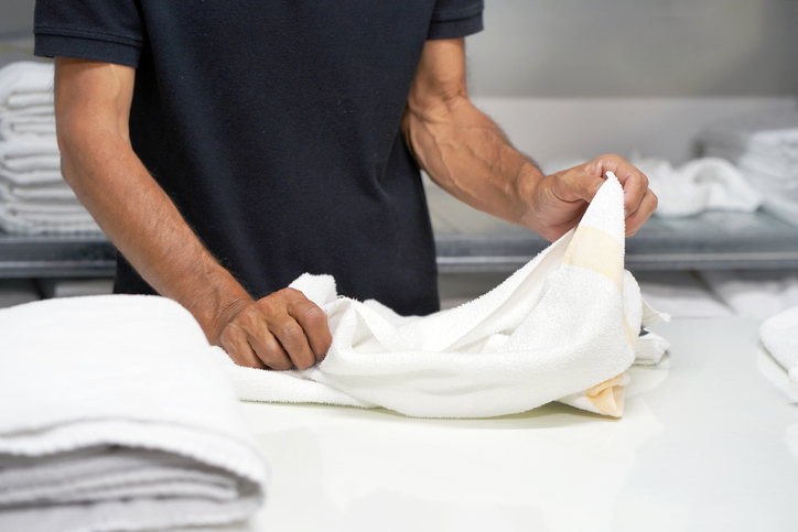 A person in a black shirt is folding a white towel on a counter, with stacks of folded towels in the background.