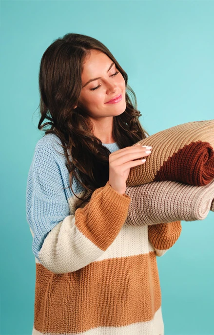 A woman in a striped sweater holds and looks at a stack of three folded knit sweaters against a plain blue background.