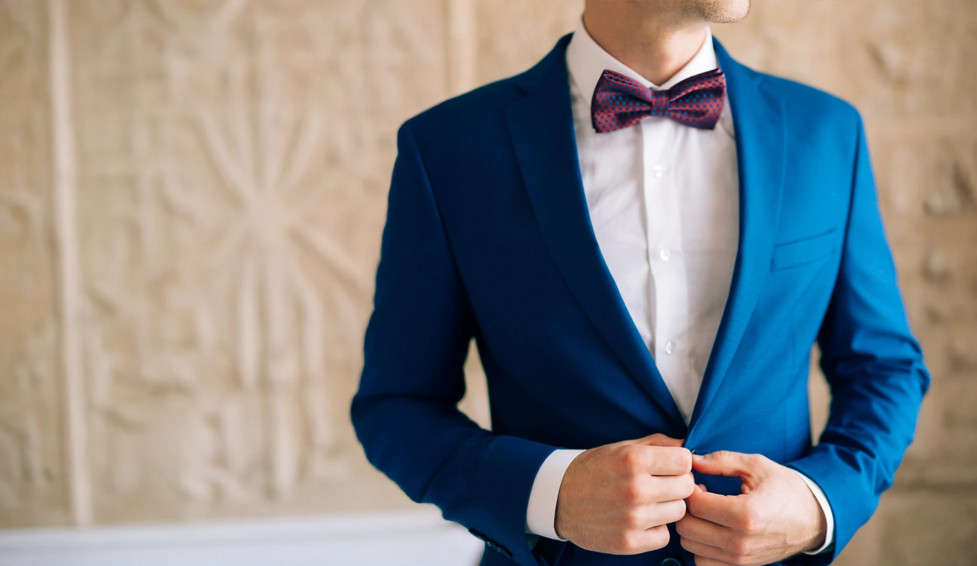A man in a blue suit and a patterned bow tie buttons his jacket. His face is partially out of frame and the background is beige with abstract designs.