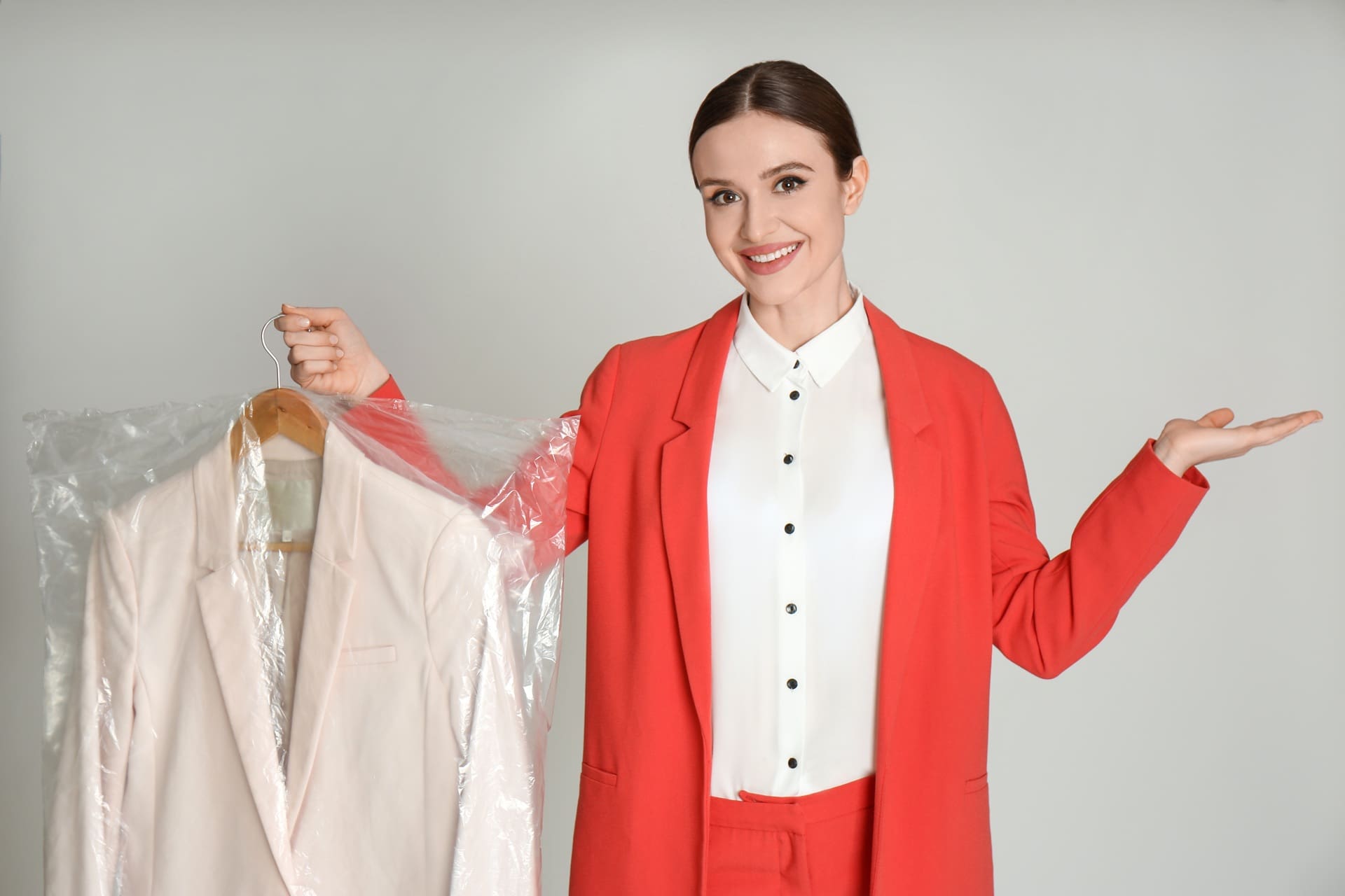 Woman in a red suit holds a clothes hanger with a light-colored jacket covered in plastic, standing against a plain background and smiling.