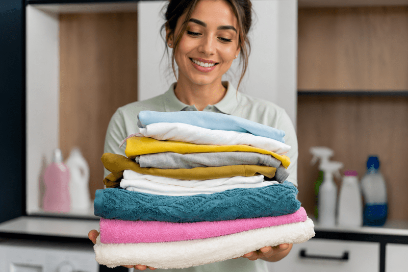 A woman smiles while holding a neatly folded stack of laundry in a modern laundry room with cleaning supplies in the background.
