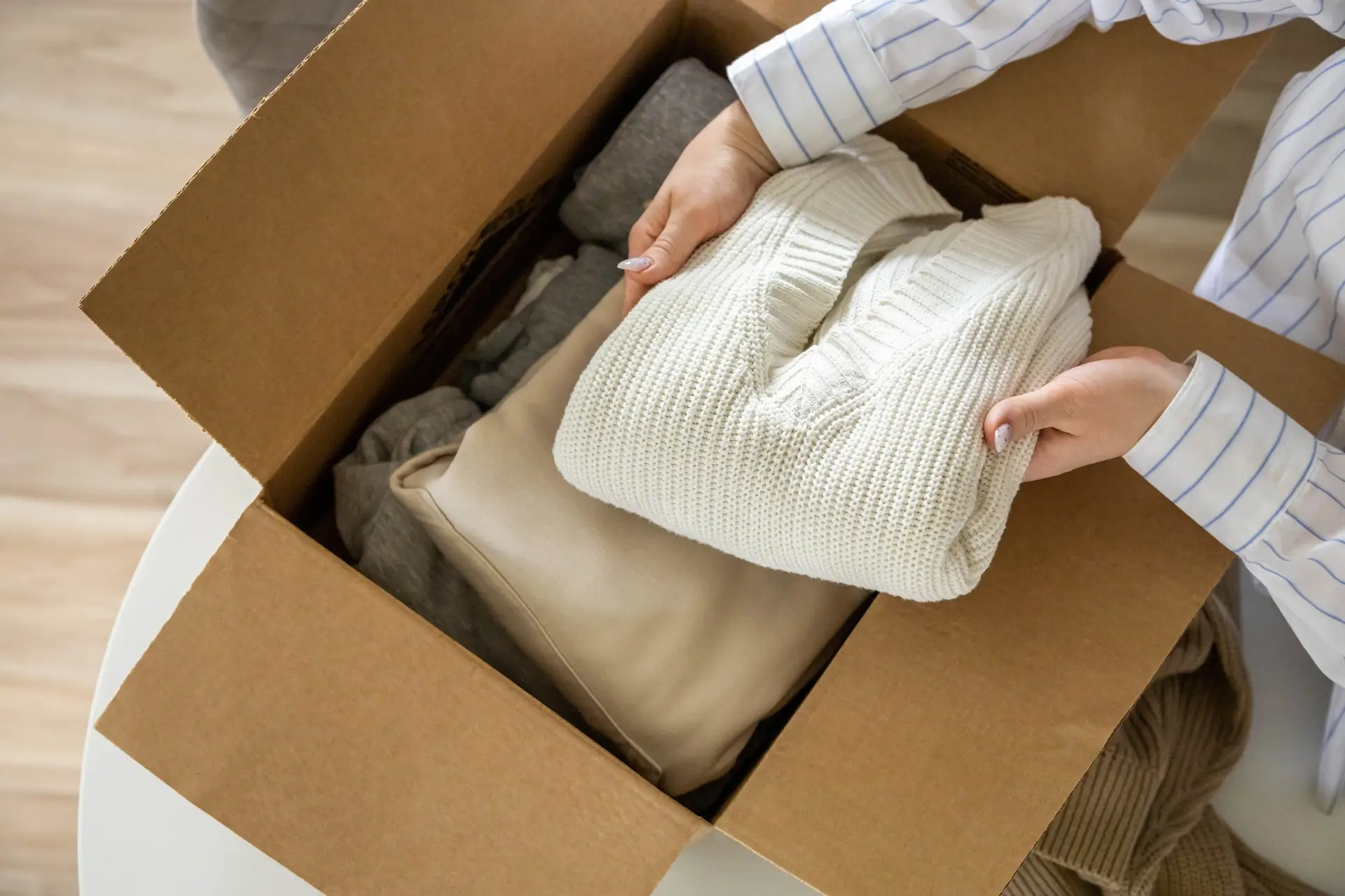 A person places a folded white sweater into a cardboard box filled with neatly packed clothing items.